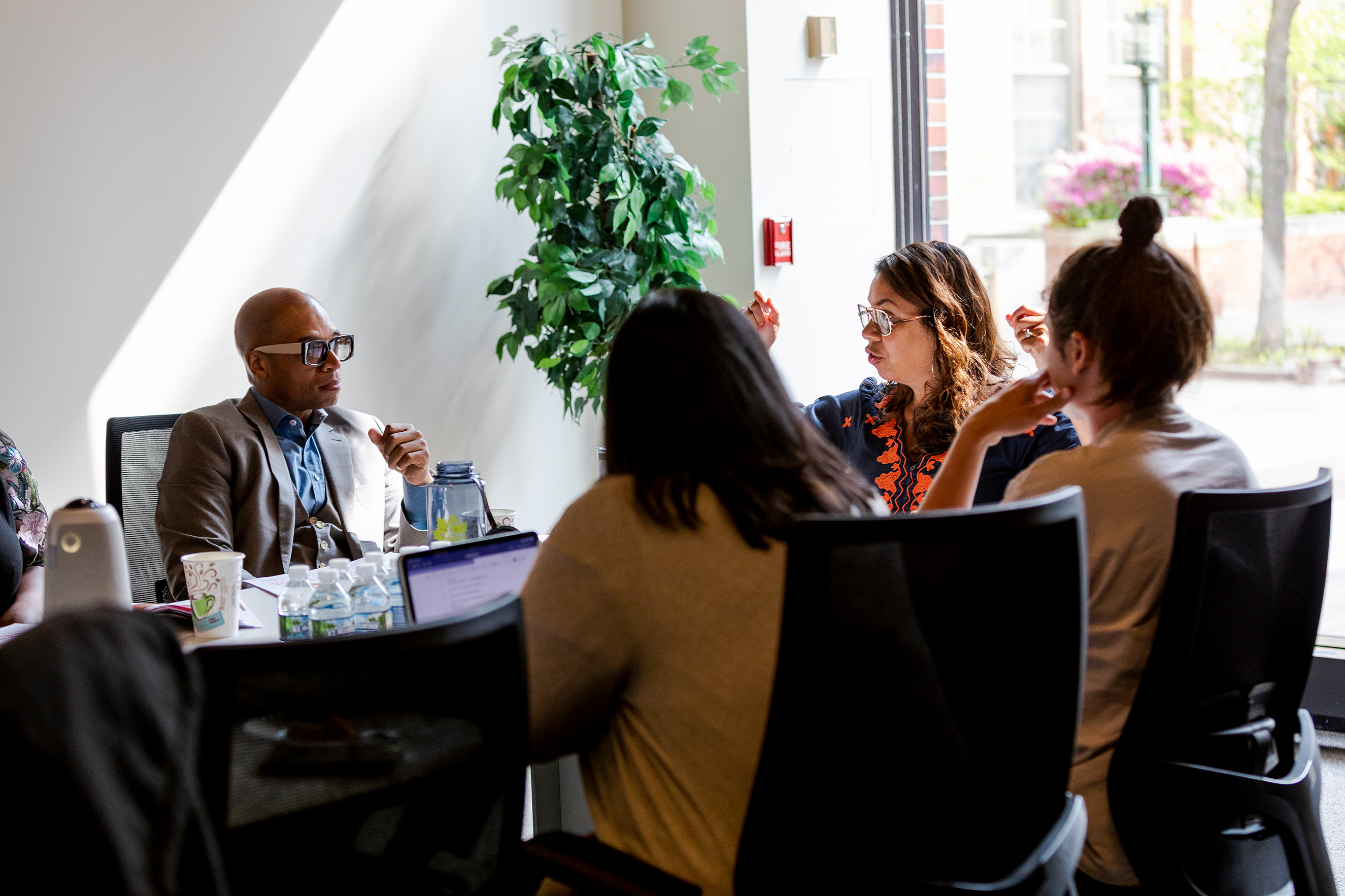 Four people sit around a table in front of a big glass window. Coffee cups, water bottles and open laptop computers are in front of them.