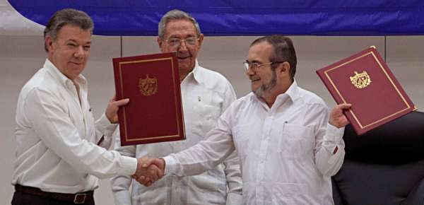 Juan Manuel Santos, Raúl Castro, and Timoleón Jimenez smile on stage and shake hands while holding red portfolios with gold trim.