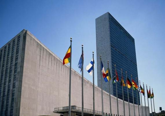 On a clear day, flags from various nations billow in the wind in front of the UN headquarters in New York: a modern rectangular skyscraper.