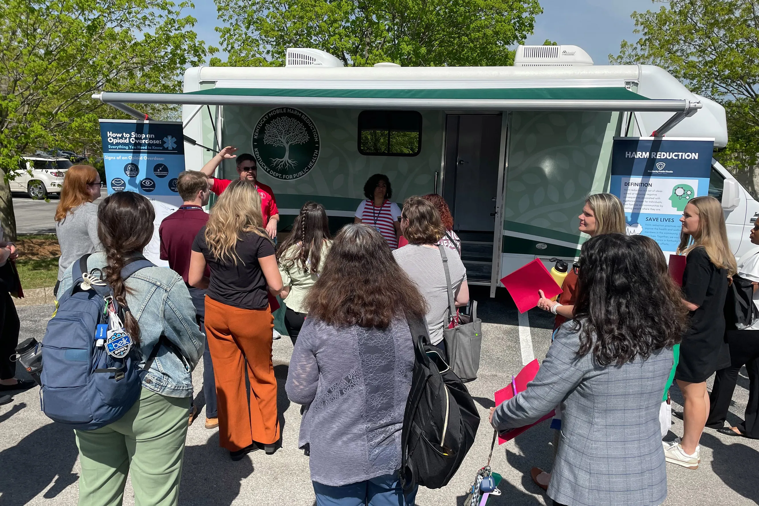 A group of people stand in front of a mobile harm reduction site with posters promoting harm reduction and opioid awareness. Trees and sunny skies are visible in the background. Some people hold red folders while listening to two individuals speaking near the clinic.