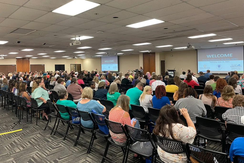 Vital Strategies is hosting a seminar on harm reduction. A large group is seated in rows in a conference room, facing a speaker and presentation screens at the front. The screens display a welcome message. The room is well-lit, with a carpeted floor and dropped ceiling.