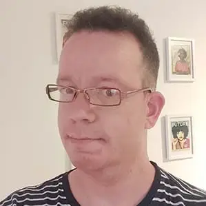 Martijn Grooten wearing a black t-shirt with white stripes against a white background with some framed images.