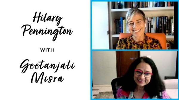 Hilary Pennington has short hair with blond highlights and is wearing a multi-colored patterned top and sitting on an orange chair. Geetanjali Misra has black shoulder length hair hair, and is wearing a flower patterned top and red and black framed eye glasses..